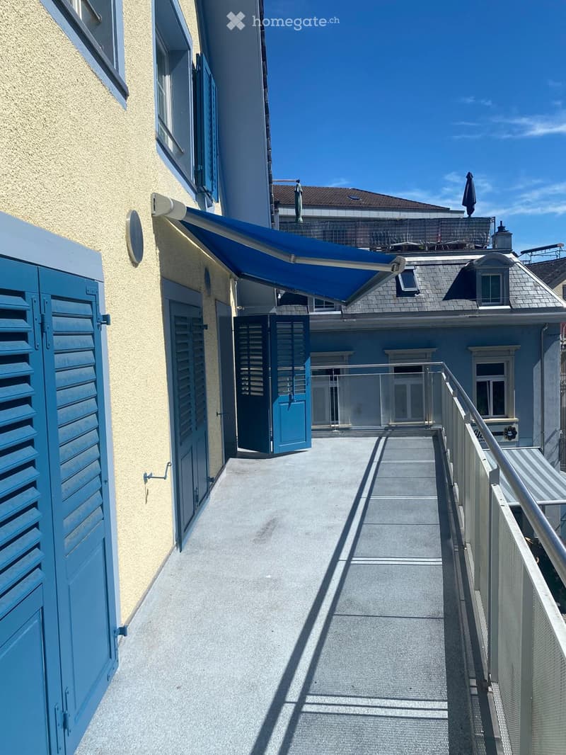 Balcony with a blue retractable awning, metal railing, and blue shutters on the adjacent building.