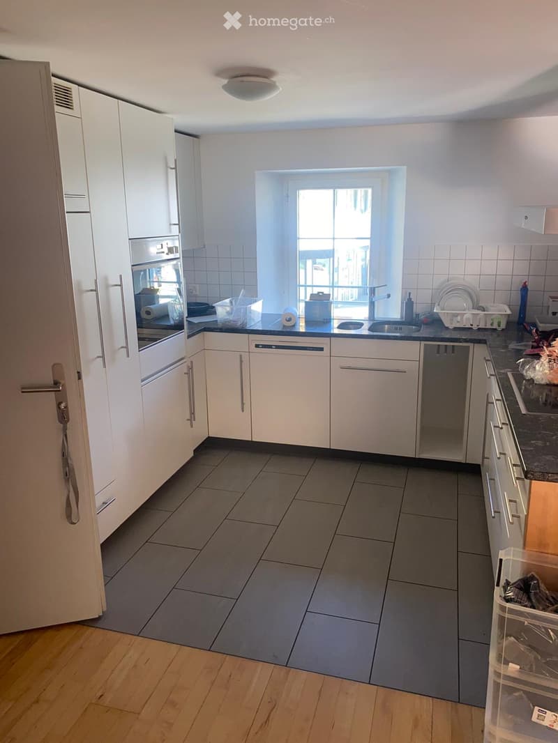 Modern kitchen with white cabinets, dark countertops, and stainless steel appliances. Features include a sink, oven, dishwasher, and a window letting in natural light.