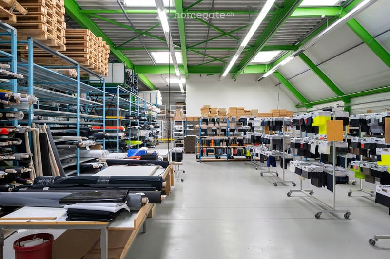 A spacious industrial warehouse with high ceilings, metal shelving units, and various materials including rolls of fabric, boxes, and workstations.