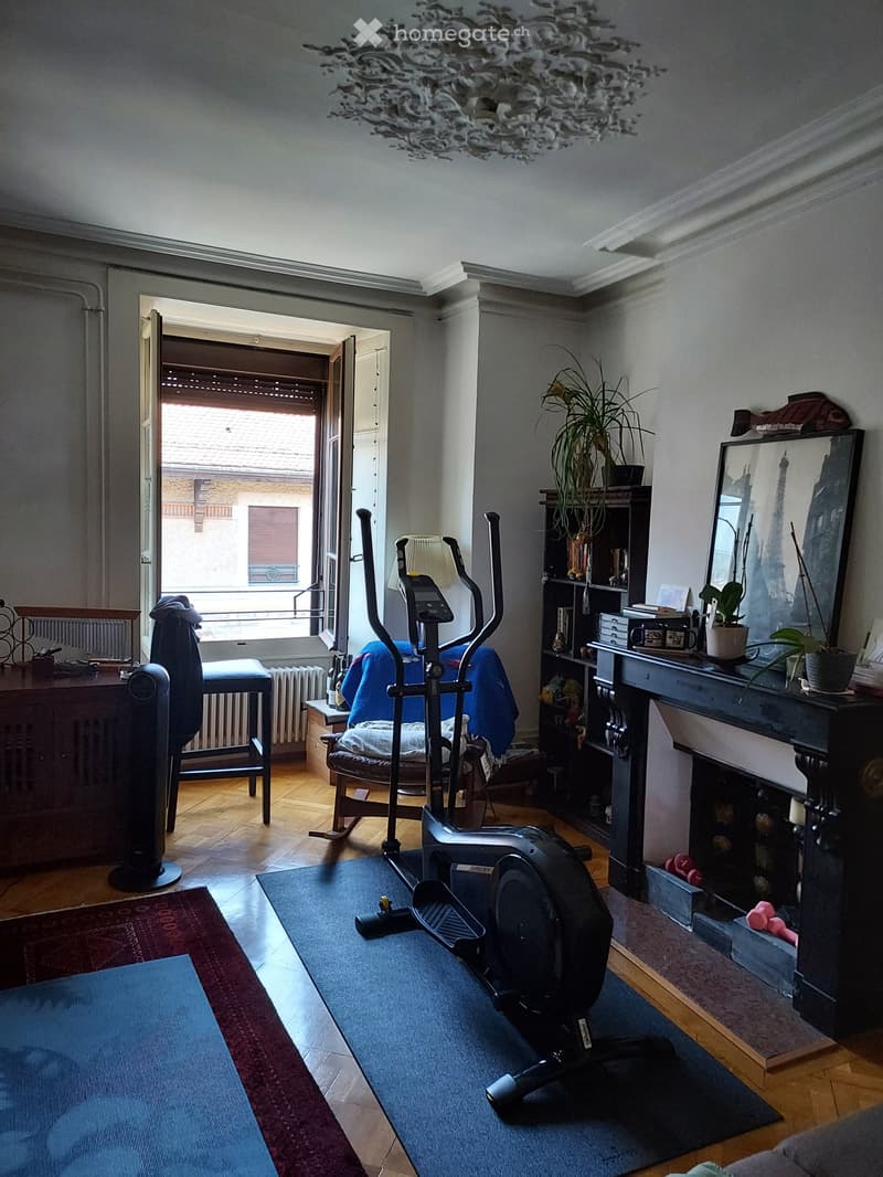 Living room with exercise bike, chair, table, mirror, plant, cabinet, fan, radiator, and a view of a balcony.
