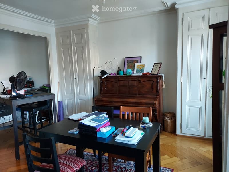 A well-furnished room with a wooden floor, a large desk with a laptop, a fan, and a piano. There are books, picture frames, and a small table with a candle on it.