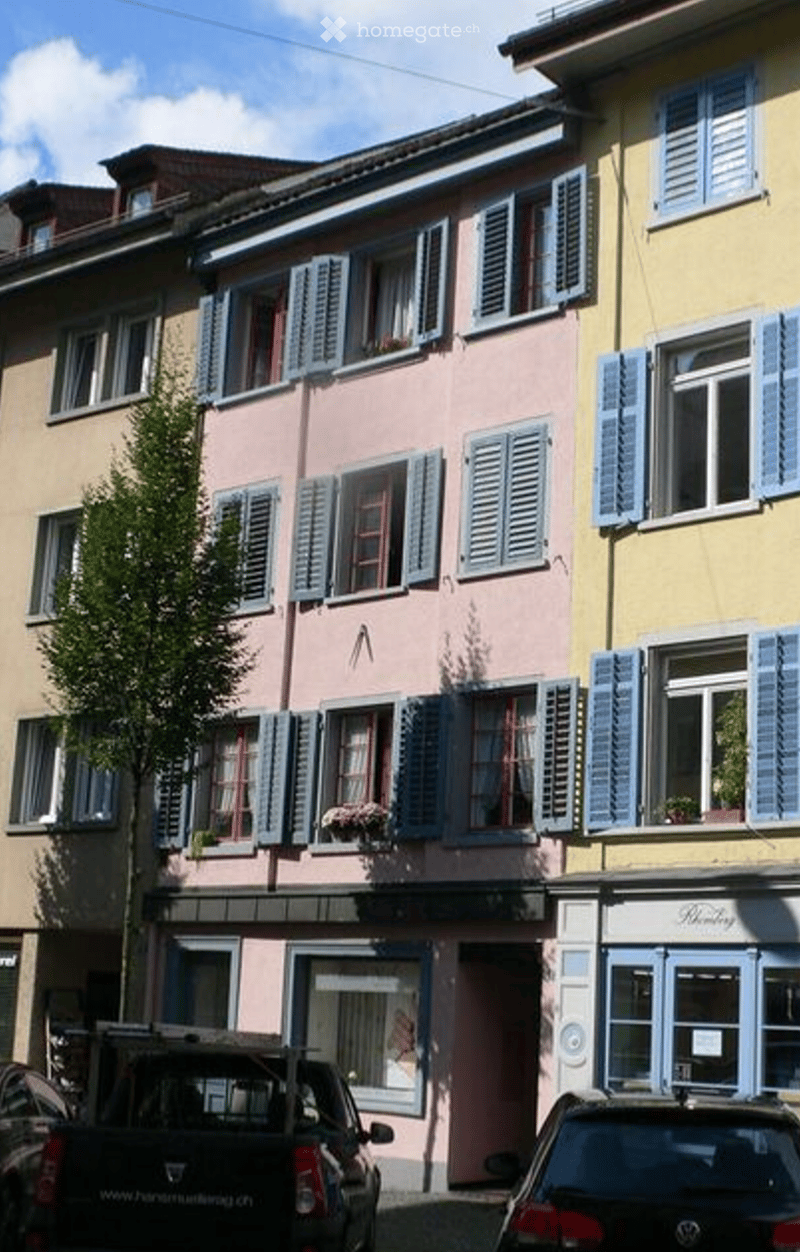 A multi-story residential building with pastel-colored facades, blue shutters, and flower boxes on the windows. The ground floor features storefronts with glass windows and doors.