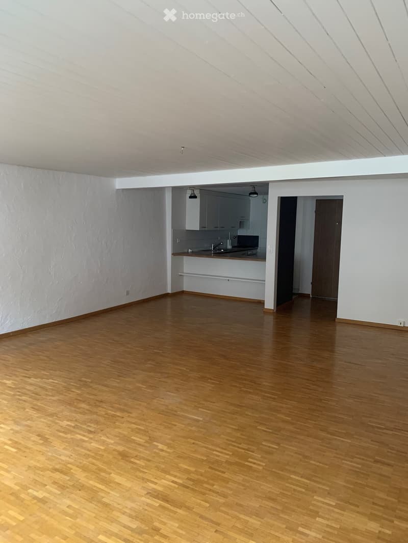 Empty room with wooden flooring, white walls, and a small kitchen area visible in the background.