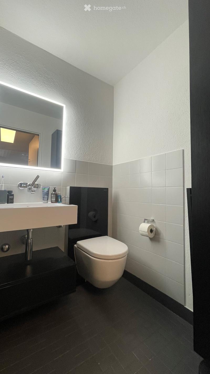 Modern bathroom with white tiles, dark floor, sink, toilet, mirror with LED lighting, and toiletries on the sink counter.