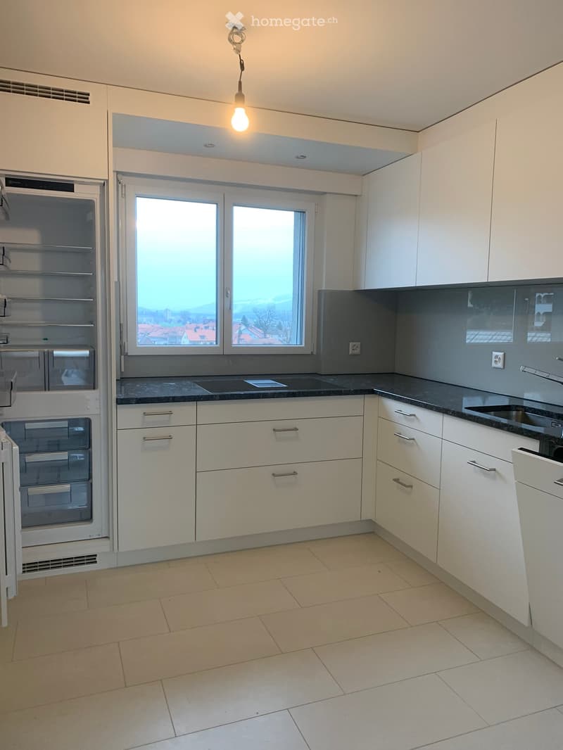 Modern kitchen with white cabinets, dark countertops, and stainless steel appliances. Features a large window with a view of rooftops.
