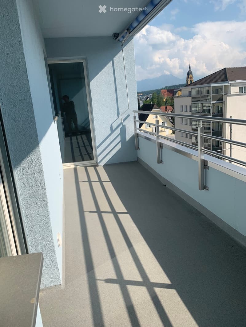 Balcony with metal railing, light-colored floor, and a view of a town with a church tower and mountains in the background.