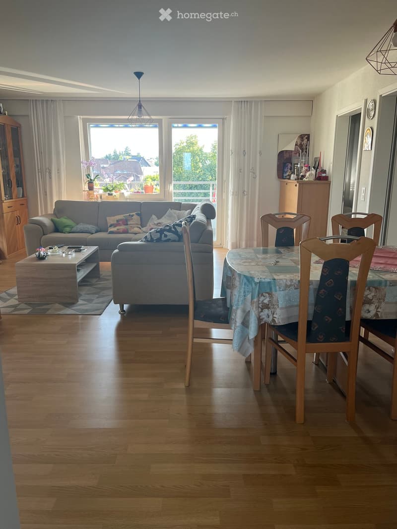 Bright living and dining area with wooden flooring, large windows with white curtains, a grey sofa with colorful cushions, a coffee table, a dining table with chairs, and a pendant light fixture.