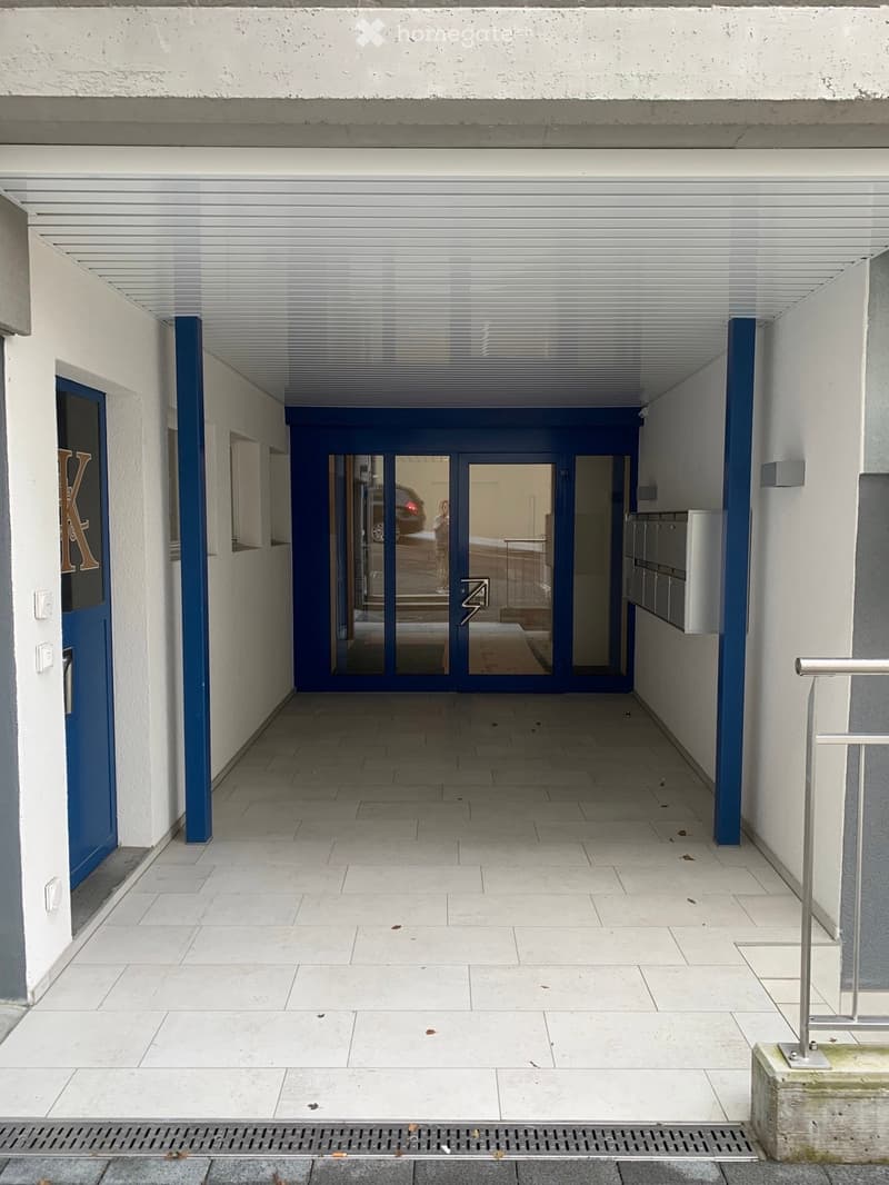 A covered entrance area with tiled flooring, white walls, and blue structural columns. Features a central glass door with blue framing leading to another area, and a side door with a blue frame and a letter "K" on it.