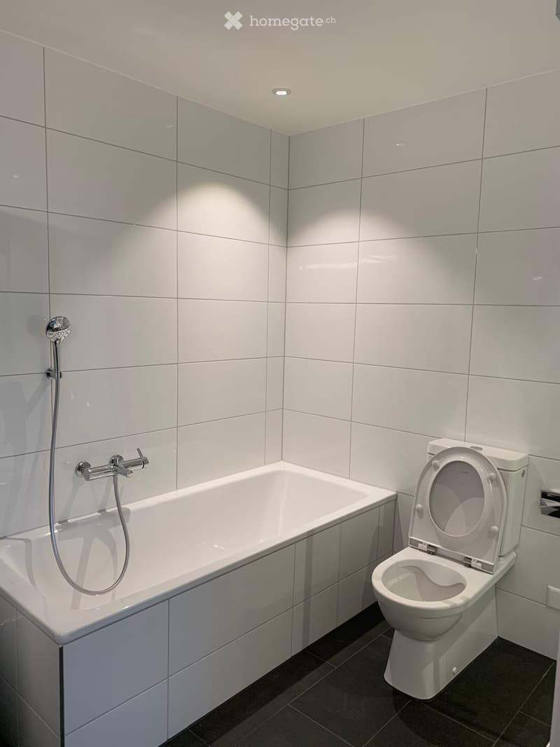 Modern bathroom with white tiled walls, a bathtub, a toilet, and a shower hose attached to the wall.