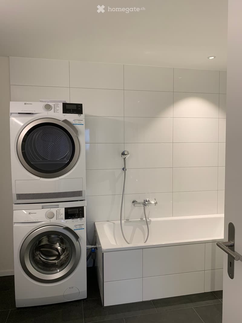 Modern bathroom with white tiled walls, a bathtub, a washing machine, and a dryer.