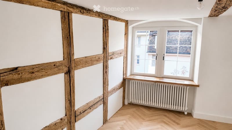 Empty room with exposed wooden beams, white walls, wooden herringbone flooring, and a large window with a radiator below.