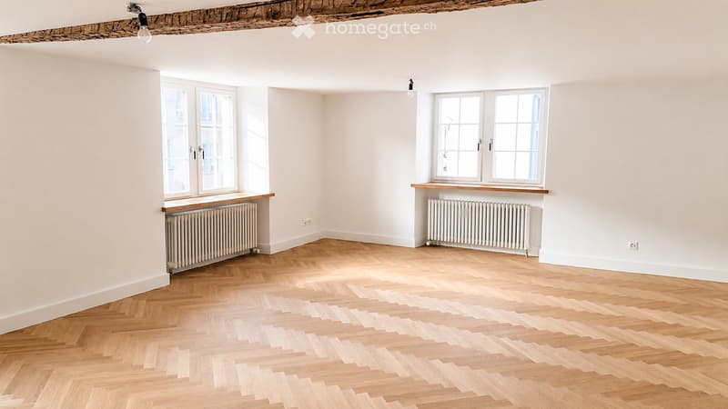 Empty room with wooden herringbone flooring, white walls, and large windows with radiators beneath them.