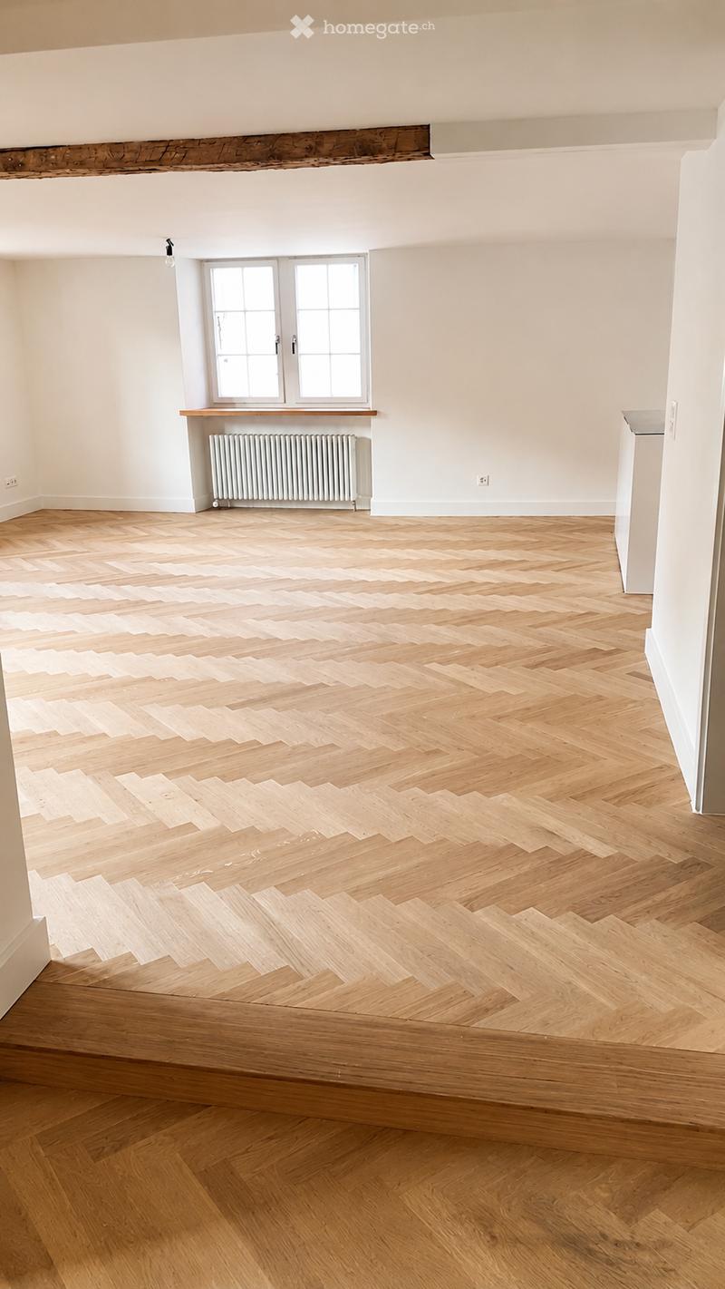 Empty room with light wooden herringbone flooring, white walls, a window with double glass panels, a radiator, and a small built-in shelf above it.