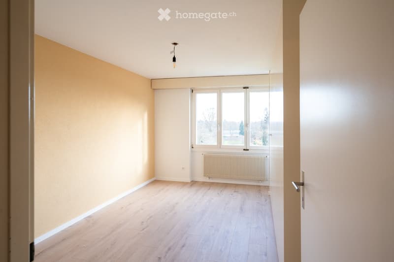 Empty room with wooden flooring, light-colored walls, large window with a view, radiator, and a single hanging light fixture.