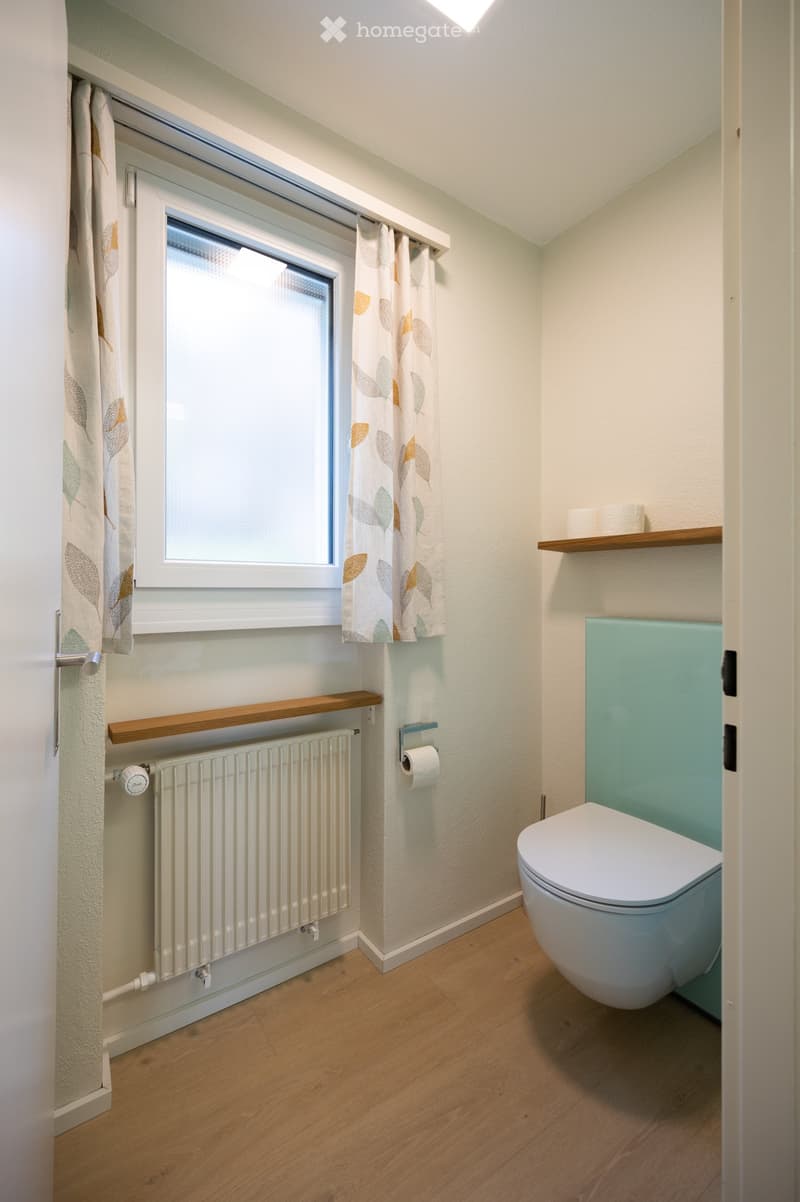 Modern bathroom with light-colored walls, wooden flooring, a toilet, radiator, and a window with a leaf-patterned curtain.
