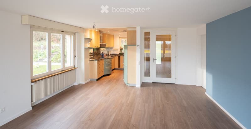 Bright, open-plan living and dining area with wooden flooring, large windows, radiator, and a modern kitchen with light wood cabinets, a stove, and a refrigerator.