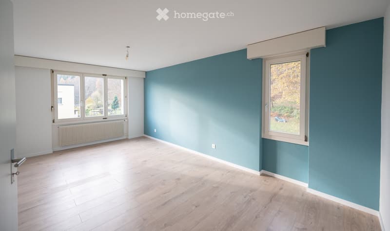 Empty room with light wooden flooring, white walls, and large windows with blue accent walls. No furniture or equipment present.