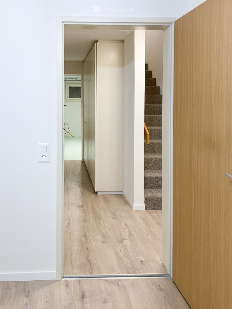 A modern, minimalist hallway with light wooden flooring, white walls, and a visible staircase leading upwards. The hallway features a glass door at the end, revealing a glimpse of a room with a washing machine and a small staircase.