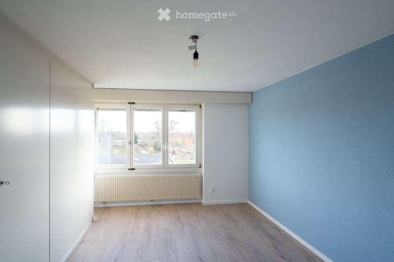 Empty room with light blue walls, wooden flooring, large windows, radiator, and a single hanging light fixture.