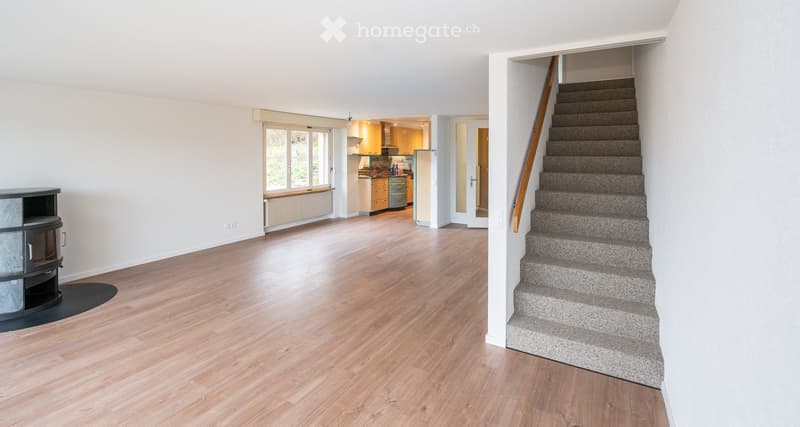 Open-plan living and dining area with wooden flooring, large windows, and a staircase leading to an upper level. The kitchen is visible in the background with cabinets and appliances.