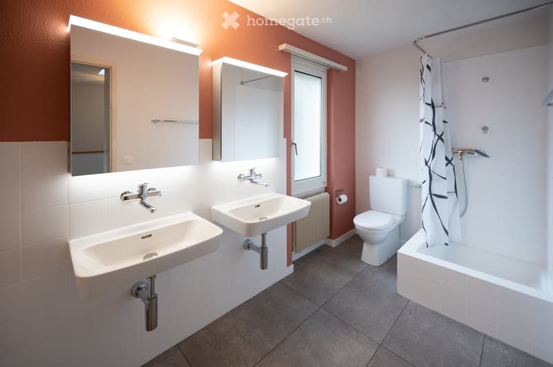 Modern bathroom with two sinks, a toilet, a bathtub, and a large mirror. The walls are painted in warm tones, and the floor is tiled.