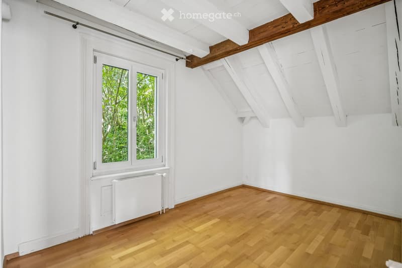Empty attic room with wooden flooring, white walls, exposed wooden beams, and a large window showing greenery outside.