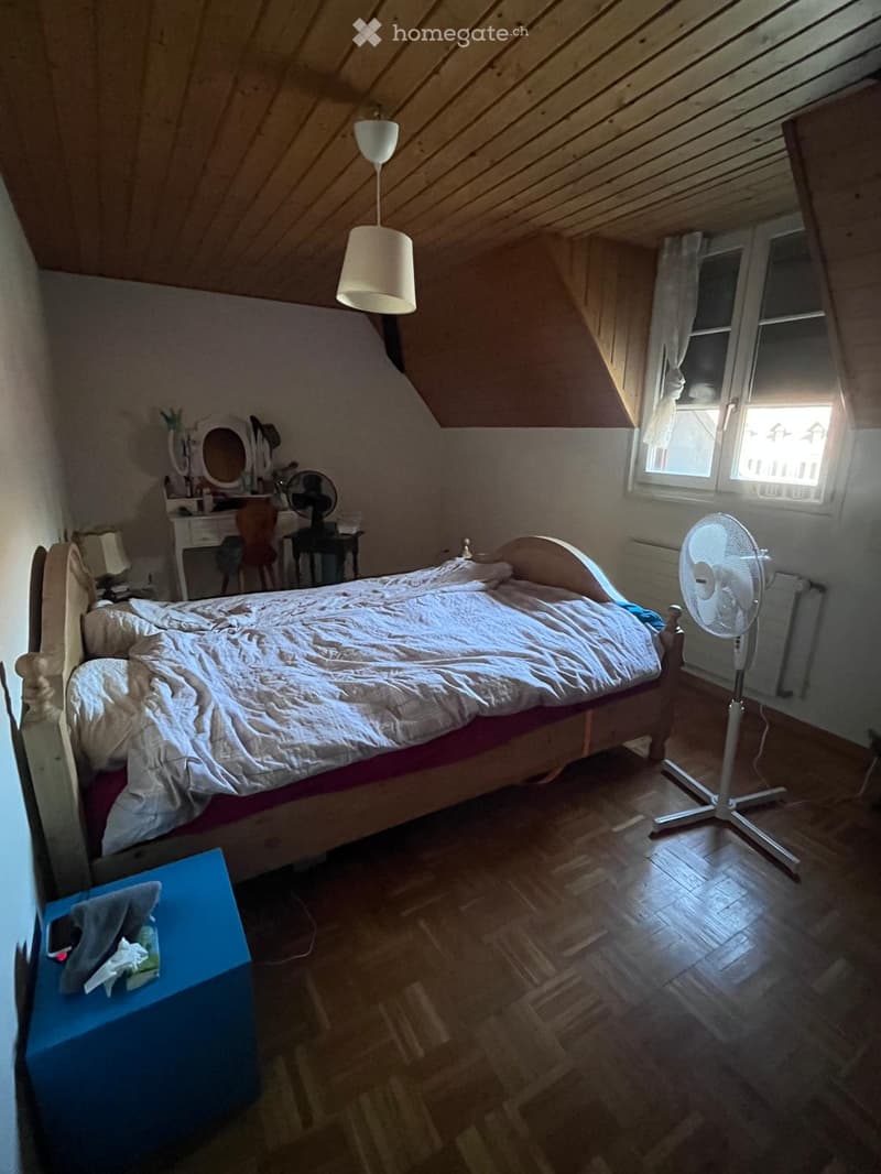 Cozy attic bedroom with wooden slanted ceilings, a large bed with white bedding, a standing fan, a small vanity with a mirror, and a hanging lamp.