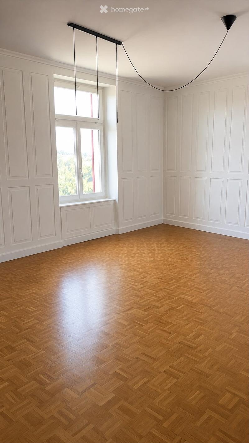Empty room with wooden herringbone flooring, white paneled walls, large windows, and a modern pendant light fixture.