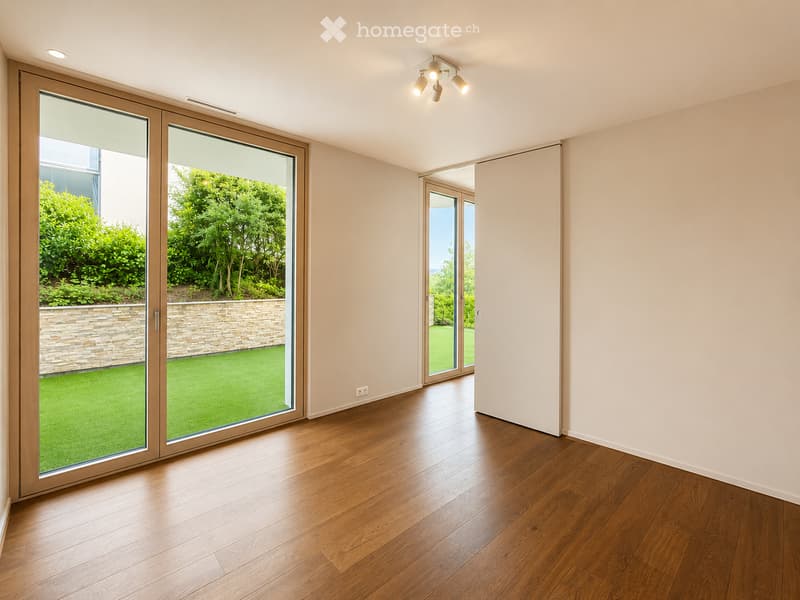 Empty room with wooden flooring, large sliding glass doors leading to a garden, and recessed ceiling lighting.