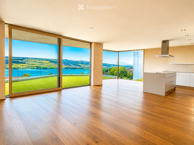 Modern, unfurnished kitchen with light wooden flooring, large panoramic windows offering scenic views, and a sleek white kitchen island.