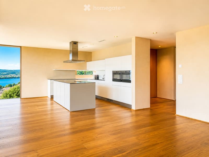 Modern kitchen with white cabinets, a central island, and a range hood, featuring wooden flooring and large windows offering a scenic view.