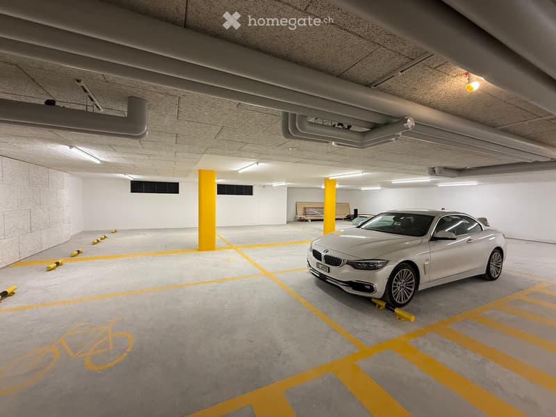 Underground parking area with a silver BMW parked in a designated spot, marked parking spaces, and yellow bollards.