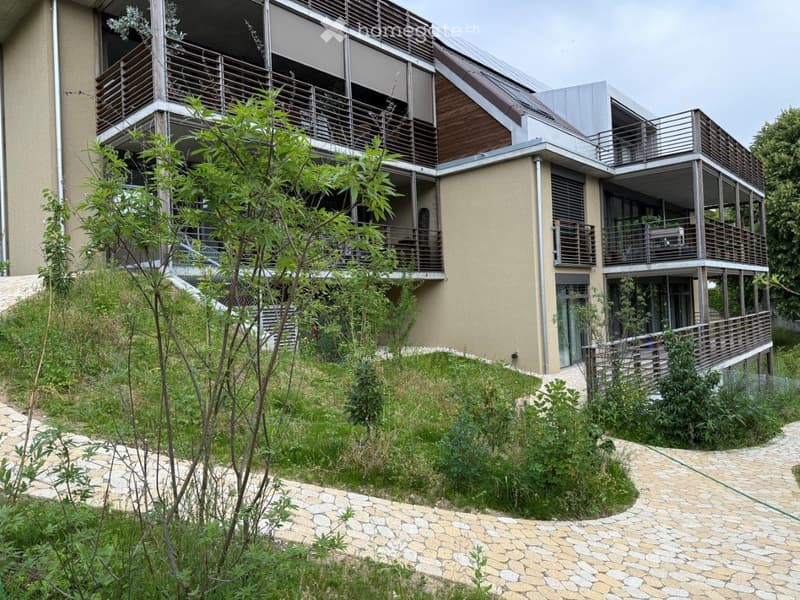 Modern two-story building with balconies, wooden railings, and a raised ground floor surrounded by greenery and a stone pathway.