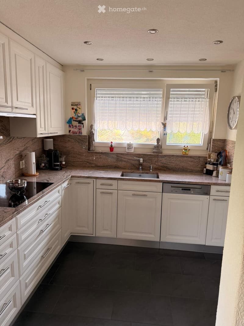 A modern kitchen with white cabinets, a central island, and a sink with a faucet. The countertops are made of marble, and the floor is tiled in dark black tiles. There is a dishwasher, a coffee maker, and a clock on the wall.