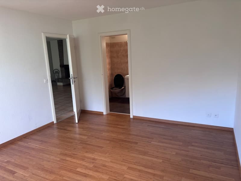 Empty room with wooden flooring and white walls, featuring two open doors leading to a bathroom and another room.