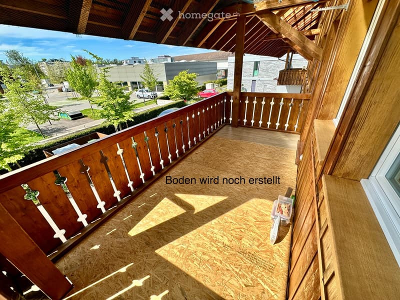 Balcony with wooden railing and unfinished wooden floor, overlooking a street with trees and buildings.