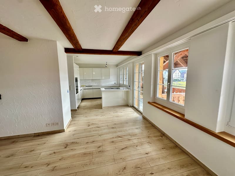 Empty room with wooden flooring, white walls, and exposed wooden beams on the ceiling. The room has large windows and a view of the outdoors.