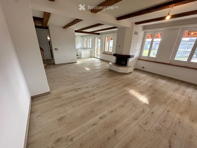 Empty room with wooden flooring, white walls, exposed wooden beams on the ceiling, and a fireplace. Large windows provide natural light, and the space is open and uncluttered.