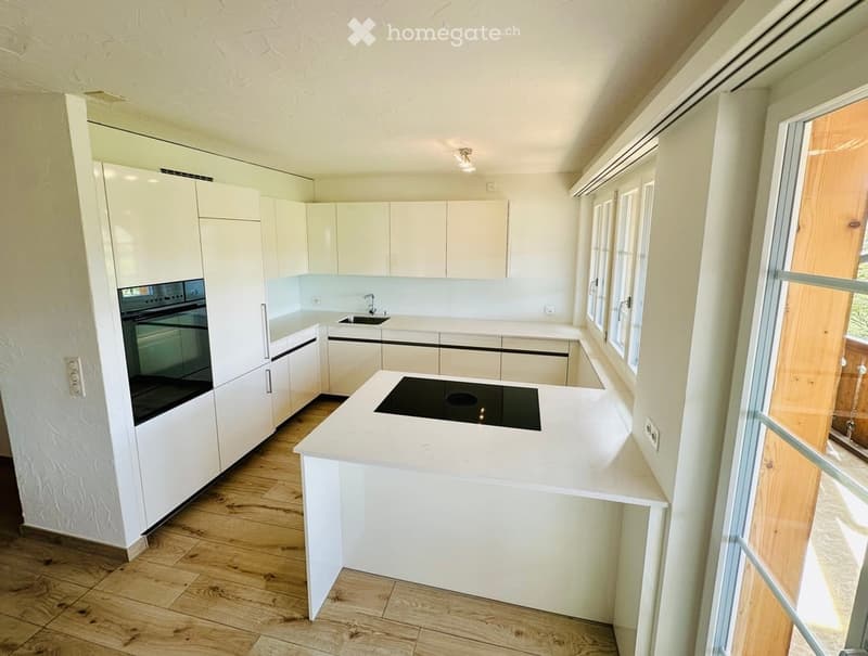 Modern kitchen with white cabinets, a central island with an induction cooktop, and a built-in oven. The space features wooden flooring and large windows allowing natural light.