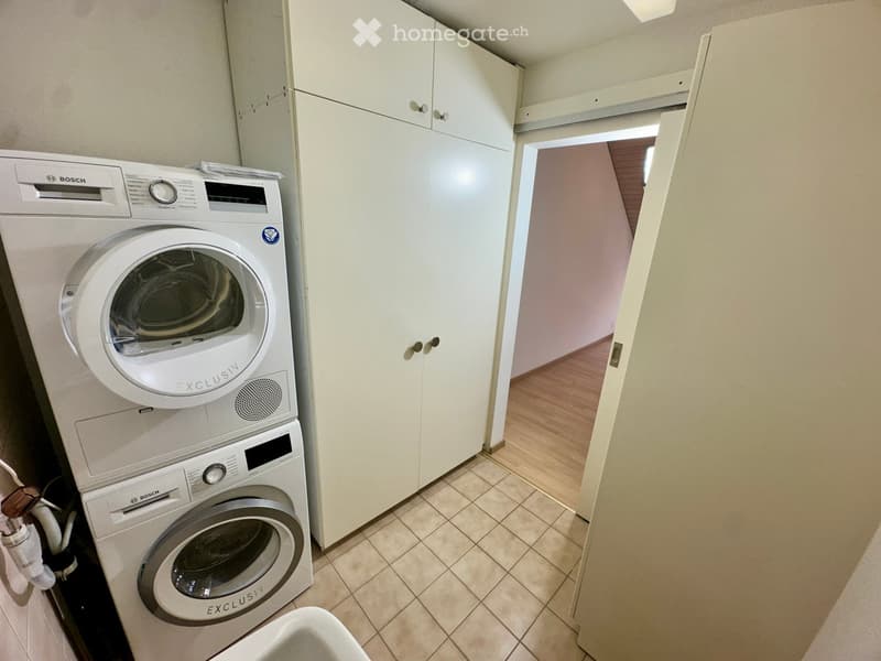 Modern bathroom with white tiled floor, white cabinets, Bosch washing machine and dryer stacked vertically, and a visible hallway beyond the open door.