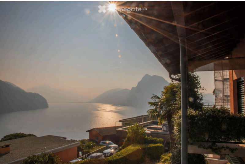 Balcony overlooking a serene lake with mountains in the background, surrounded by greenery and parked cars below.