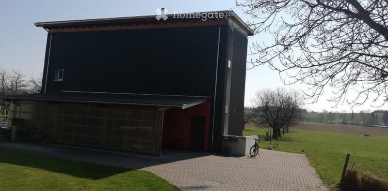 Modern two-story building with dark grey exterior, red garage section, and a paved driveway leading to a grassy area.