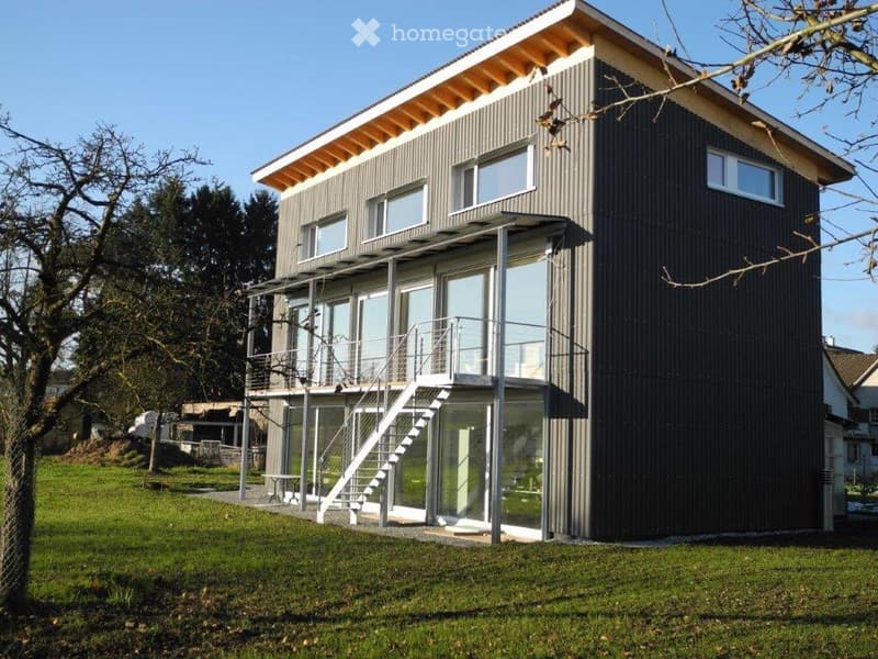 Modern two-story house with dark grey exterior, large windows, and a balcony accessible from the upper floor.