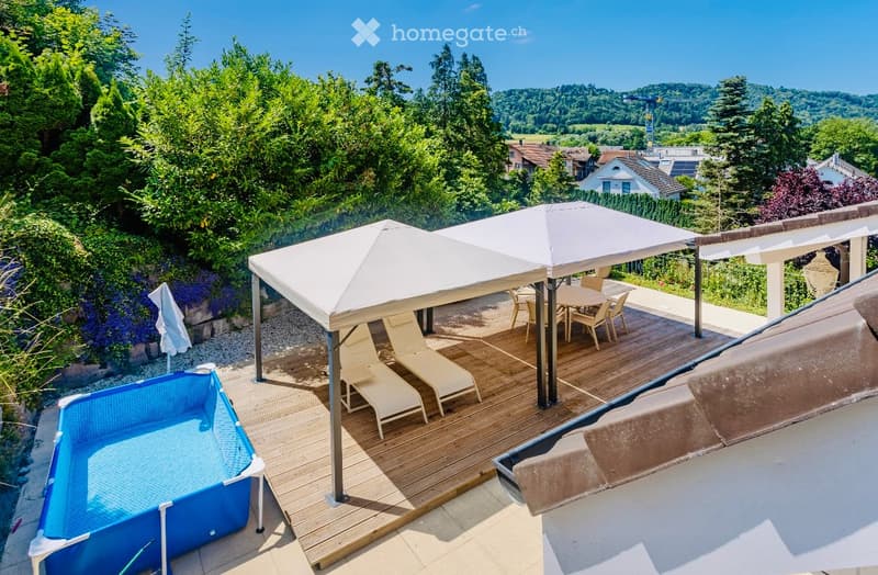 wooden deck with canopies and loungers, small rectangular pool, table and chairs under canopies, rooftop, trees, bushes, and distant houses