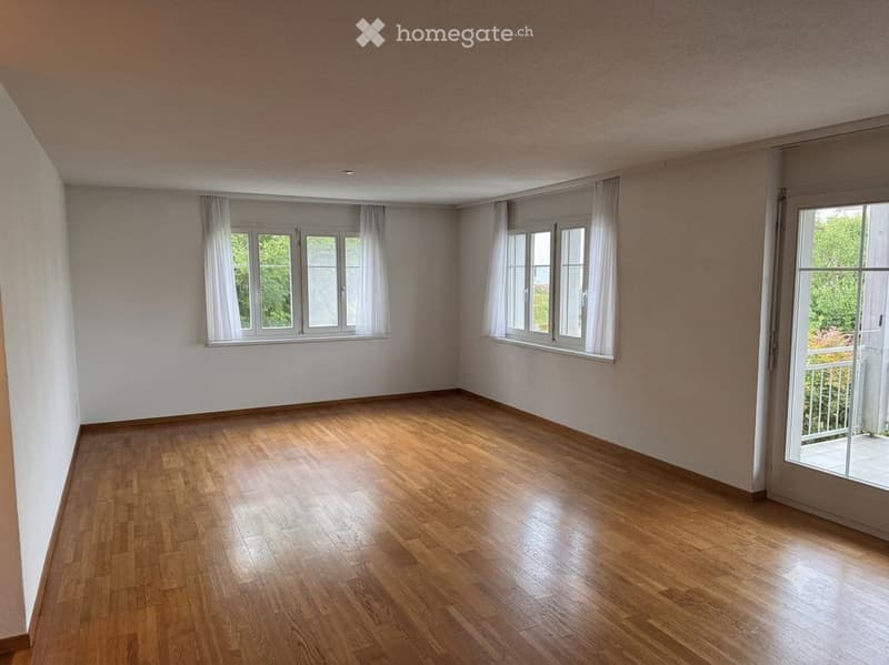 Empty room with wooden flooring, white walls, large windows with white curtains, and a sliding glass door leading to a balcony.
