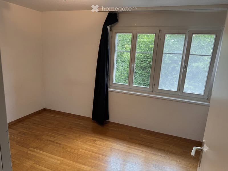 Empty room with wooden flooring, large window with black curtain, and white walls.