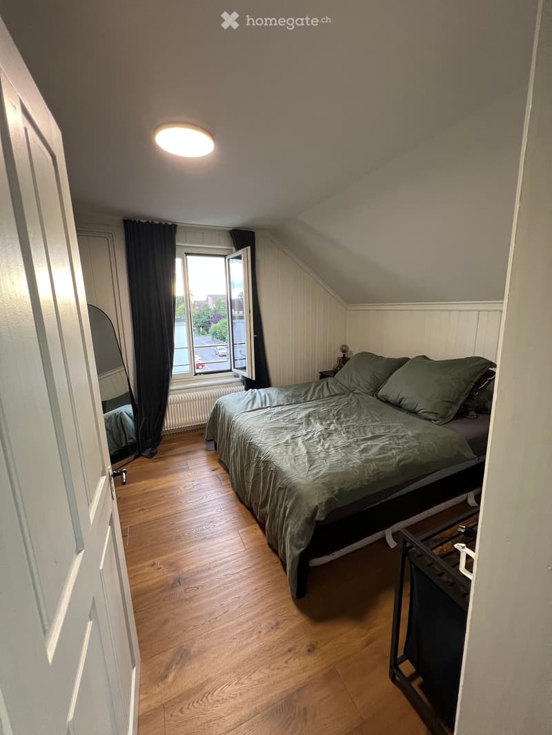 Cozy attic bedroom with wooden flooring, a double bed with green bedding, a nightstand, and a large window with dark curtains.