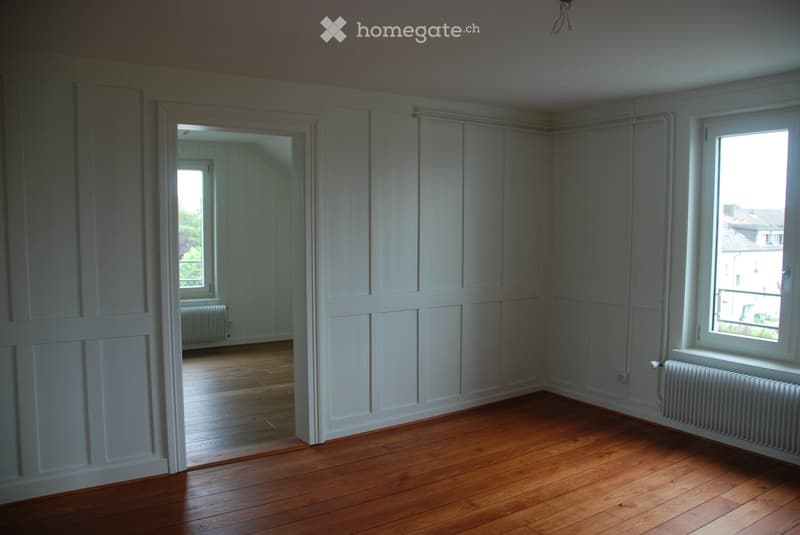 Empty room with wooden flooring, white walls, and large windows. The room is unfurnished, with visible radiators and a doorway leading to another room.