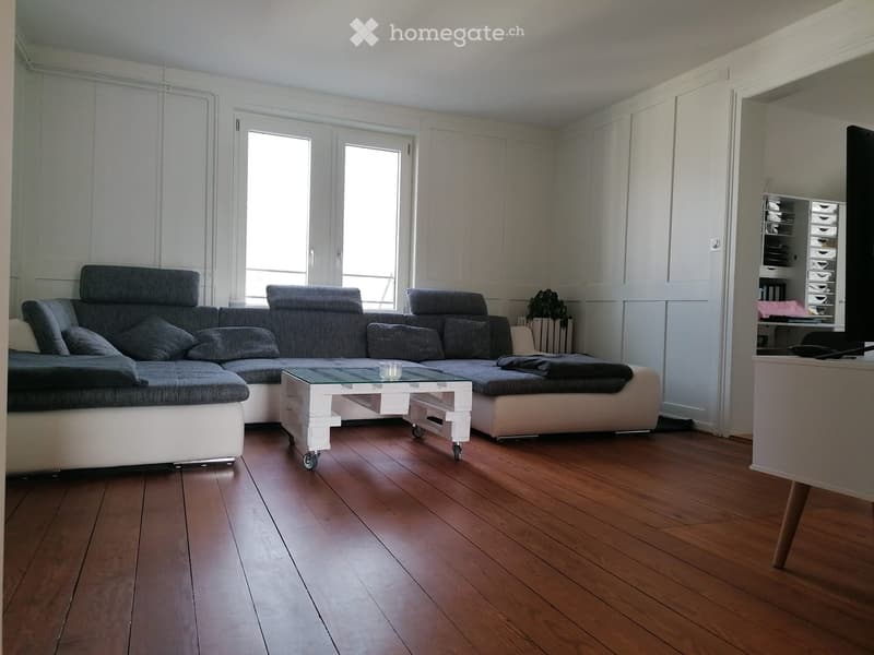 Modern living room with a grey sectional sofa, white coffee table, and a TV on a stand. The room features wooden flooring and large windows.