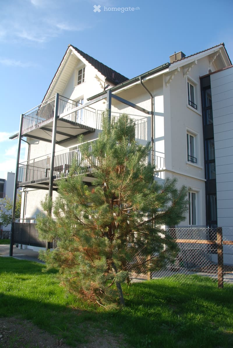 Two-story house with white exterior, multiple windows, and a balcony on the upper level. A small evergreen tree is in the front yard, enclosed by a chain-link fence.